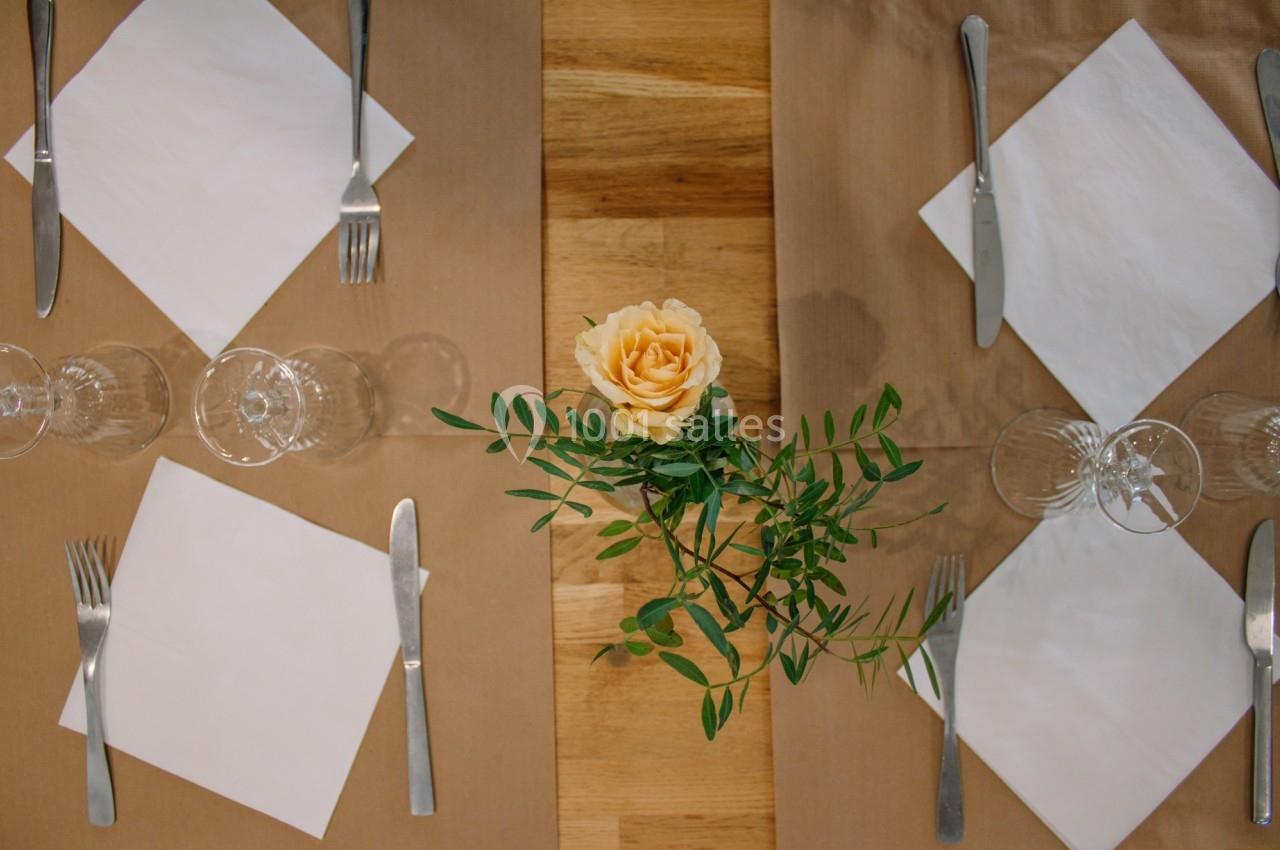 Table dressée avec des sets marron, serviettes blanches pliées, couverts, verres et une rose jaune au centre.