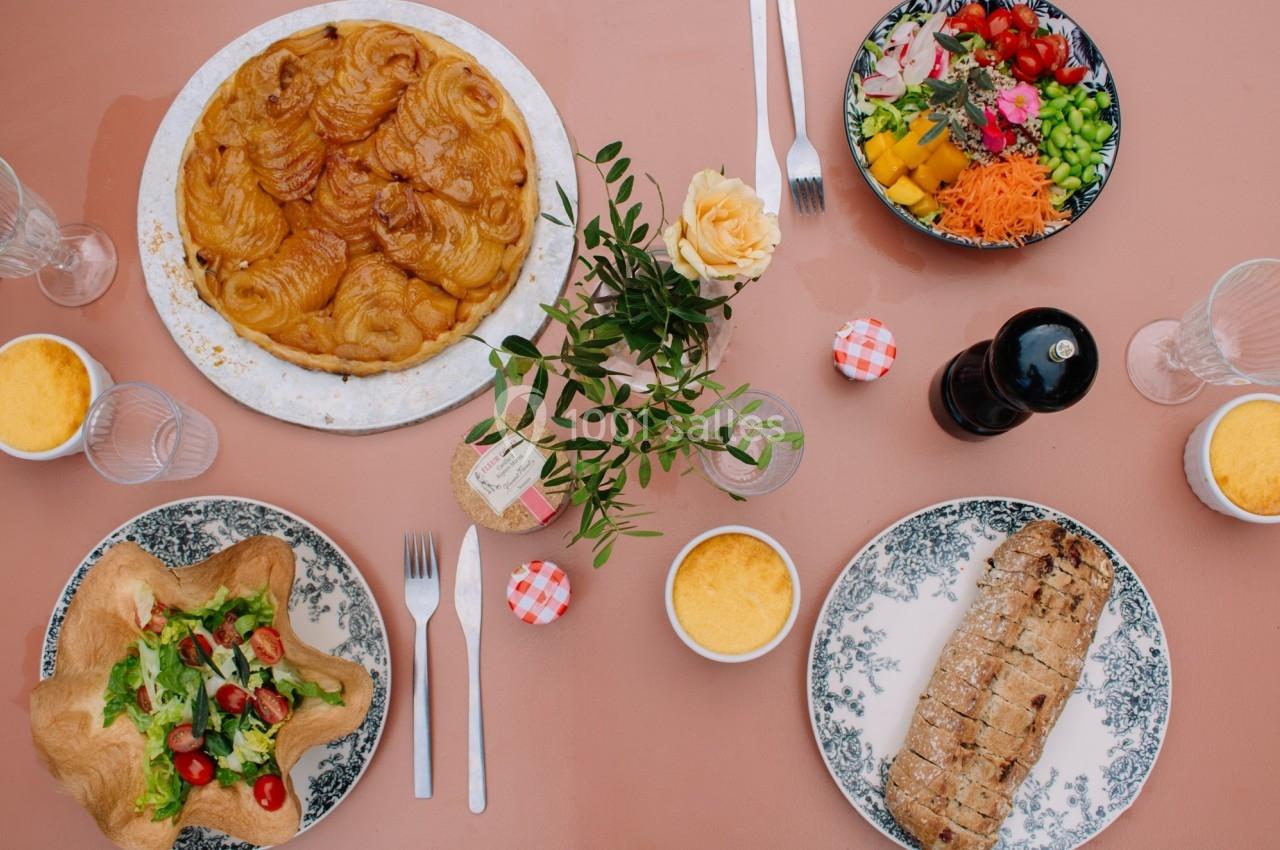 Table dressée avec une tarte aux pommes, une salade, du pain, des verrines et des couverts sur une nappe rose.