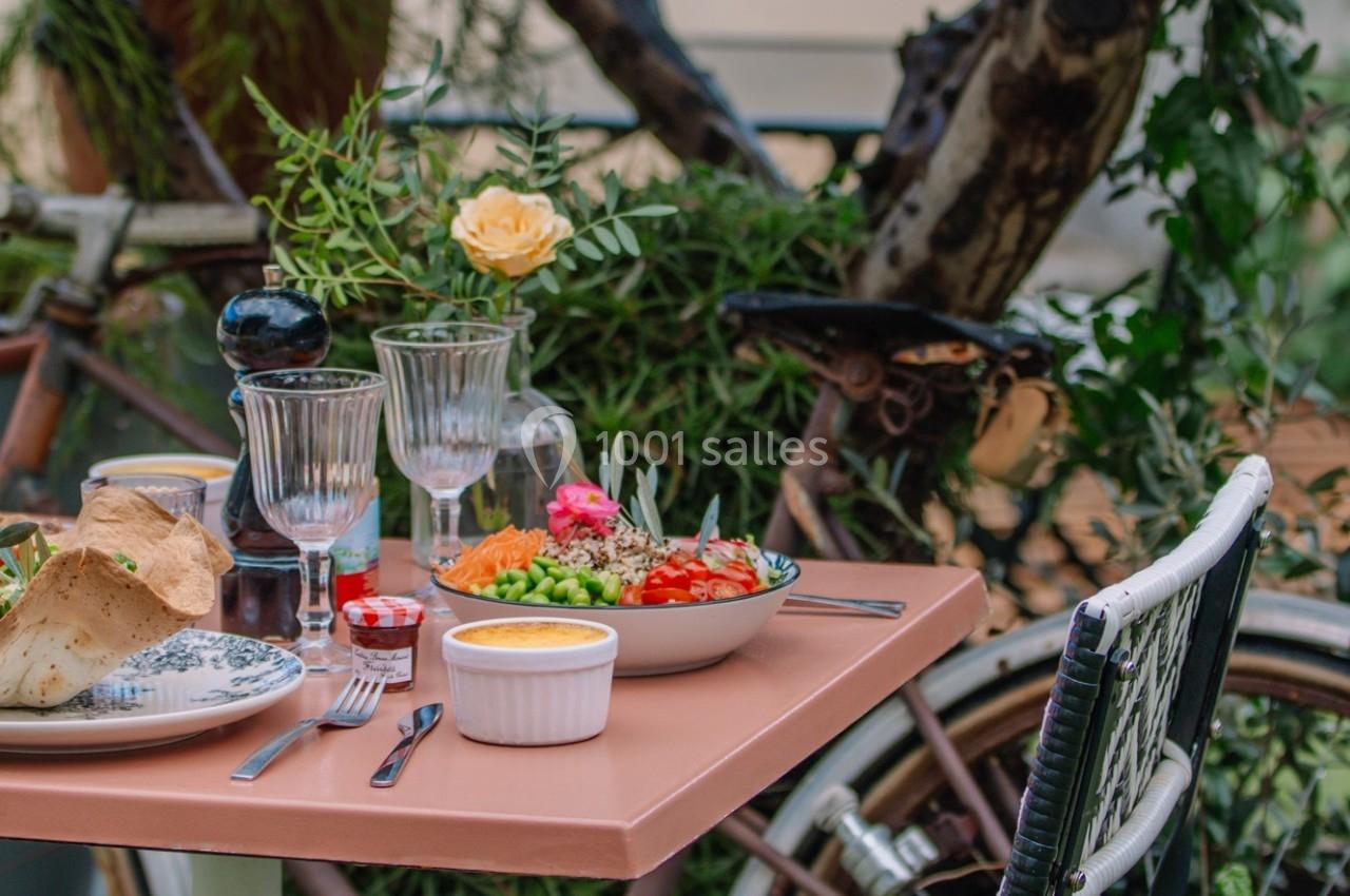 Table dressée en extérieur avec des plats colorés, verres, couverts et une décoration végétale en arrière-plan.