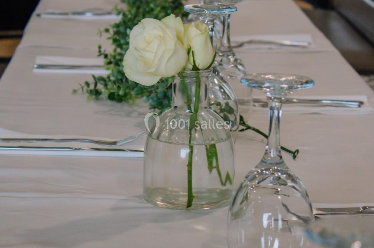 Vase en verre contenant deux roses blanches, posé sur une table dressée avec des verres et des couverts.