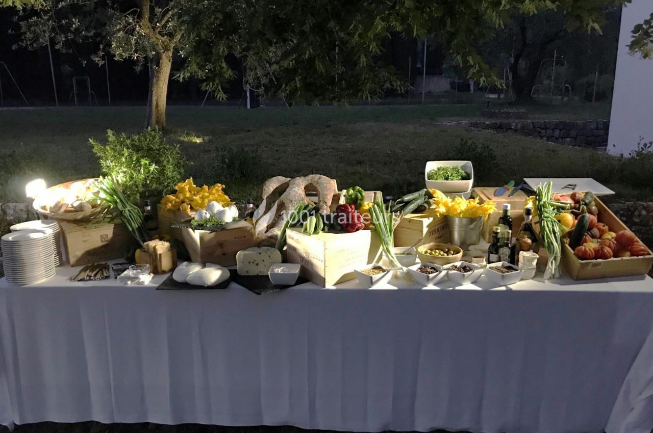Table de buffet en extérieur avec légumes frais, fromages, pains et condiments, éclairée sous des arbres.