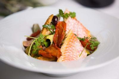Assiette de filets de poisson accompagnés de légumes rôtis, herbes fraîches et olives, présentée sur une table blanche.