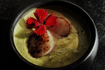 Velouté vert garni de deux noix de Saint-Jacques poêlées et décoré d'une fleur rouge, présenté dans un bol noir.
