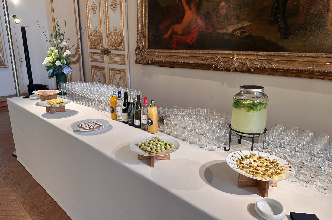 Buffet élégant avec amuse-bouches, boissons variées et décoration florale dans une salle ornée de tableaux et moulures…