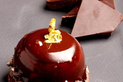 Dessert au chocolat brillant décoré de feuille d'or, accompagné de morceaux de chocolat sur une surface sombre.