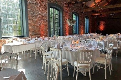 Salle de réception avec tables rondes dressées, chaises blanches et murs en briques apparentes sous un toit en poutres.