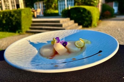 Assiette de coquilles Saint-Jacques dressées avec des fleurs comestibles et une sauce, en extérieur près d'un bâtiment.