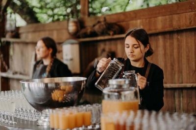 Une femme verse une boisson dans un verre sur un comptoir avec des jus et des verres alignés, en arrière-plan une autre…