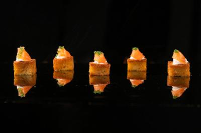 Cinq bouchées apéritives alignées, composées de pain doré, saumon fumé et garniture verte, sur fond noir.