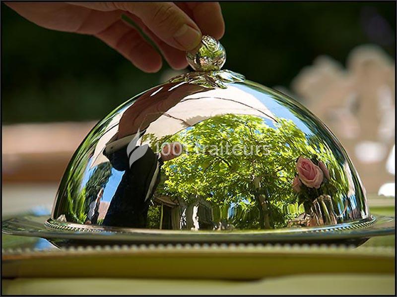 Une main soulève un couvercle en métal brillant, reflétant un arbre et des éléments environnants.