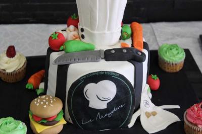 Gâteau décoré en forme de toque et tablier de chef, entouré de légumes, cupcakes et accessoires culinaires.