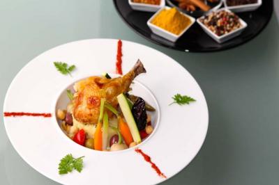 Plat de couscous garni de légumes, pois chiches et cuisse de poulet, avec épices en arrière-plan.