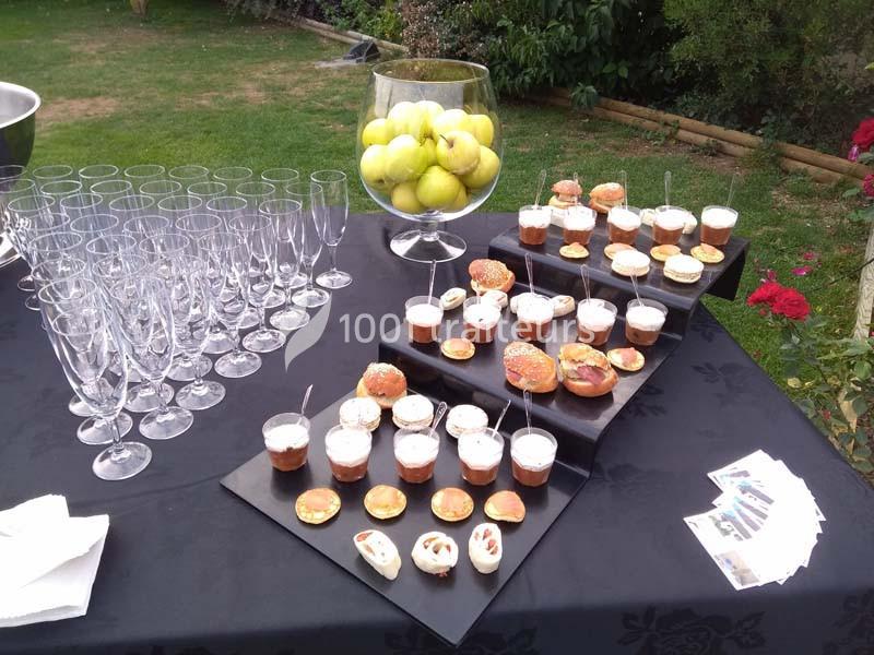 Table dressée avec des verrines, amuse-bouches, verres vides et un grand vase rempli de pommes vertes, en extérieur.