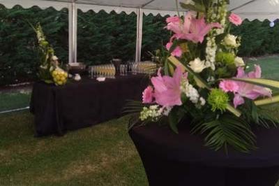 Arrangement floral avec des lys roses sur une table haute, devant un buffet sous une tente blanche en extérieur.