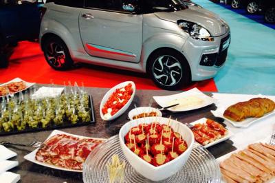 Buffet varié avec charcuterie, tapas et viennoiseries en premier plan, petite voiture grise exposée en arrière-plan.