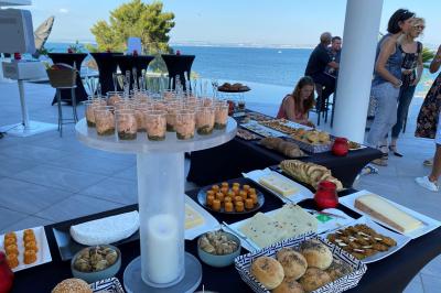 Buffet en extérieur avec vue sur la mer, comprenant pains, viennoiseries, fromages et verrines disposés sur des tables.