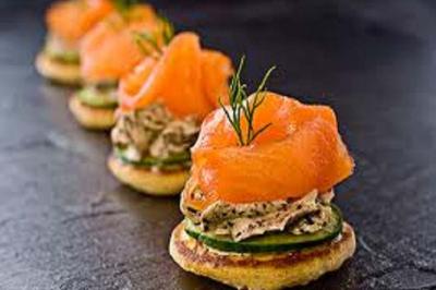 Blinis garnis de saumon fumé, crème aux herbes, rondelles de concombre et brins d'aneth, alignés sur une surface sombre.
