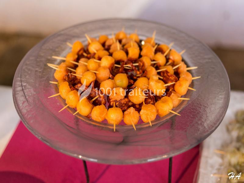 Assiette de brochettes apéritives composées de billes de melon et de morceaux de charcuterie, présentée sur une table.