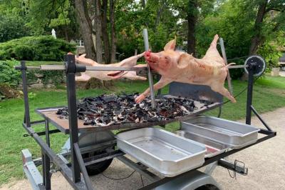 Cochon entier en cours de cuisson sur une broche rotative au-dessus de braises, dans un cadre extérieur verdoyant.