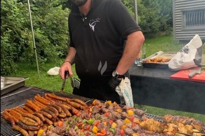 Homme cuisinant des brochettes et saucisses sur un grand barbecue en plein air, avec un jardin en arrière-plan.