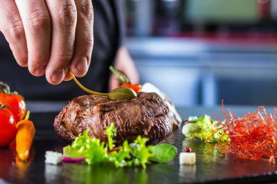 Une main dépose une sauce sur un steak accompagné de légumes et de garnitures colorées sur une assiette noire.