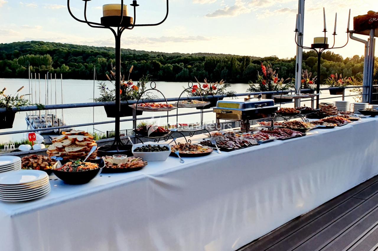 Buffet en plein air avec divers plats et assiettes, installé sur une terrasse surplombant un lac au coucher du soleil.