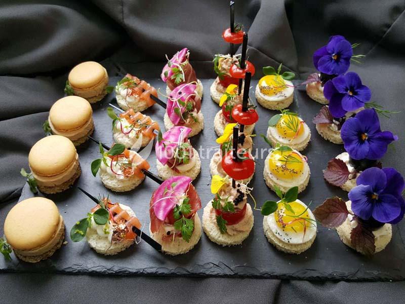 Plateau de canapés variés avec macarons salés, légumes, fleurs comestibles et décorations colorées sur ardoise.