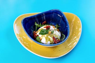 Assiette colorée contenant une burrata garnie de pesto, tomates séchées, pignons de pin et roquette sur fond bleu.