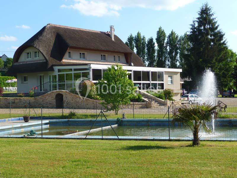 Maison au toit de chaume avec terrasse vitrée, entourée de verdure, d'un bassin et d'une fontaine en premier plan.