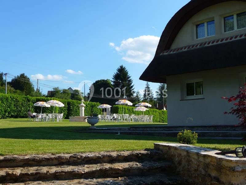 Jardin avec pelouse, tables et parasols colorés, entouré de haies, près d'une maison au toit arrondi.