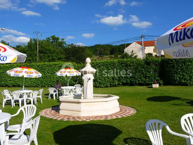 Fontaine centrale entourée de tables et chaises blanches sur une pelouse, avec des parasols colorés sous un ciel bleu.