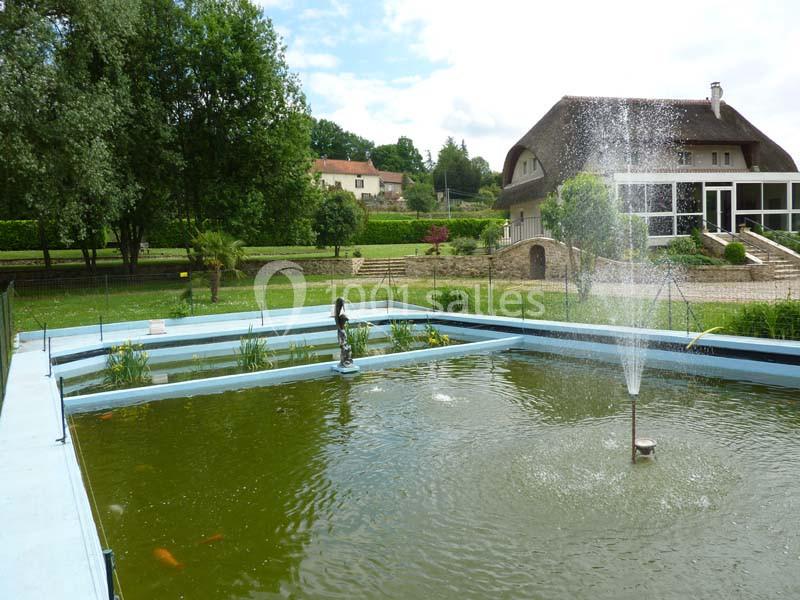 Bassin extérieur avec fontaine centrale, poissons visibles et végétation environnante, maison en arrière-plan.