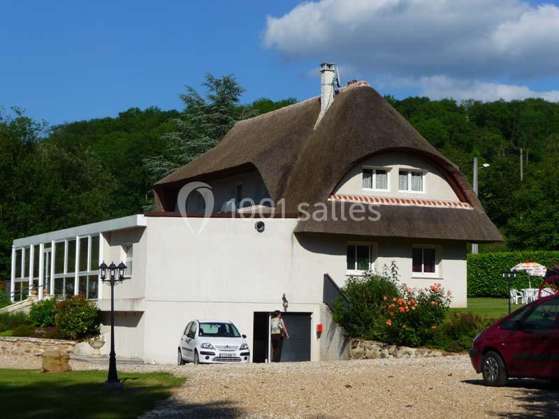 Maison avec toit de chaume, garage ouvert, jardin fleuri et allée gravillonnée, entourée de verdure.
