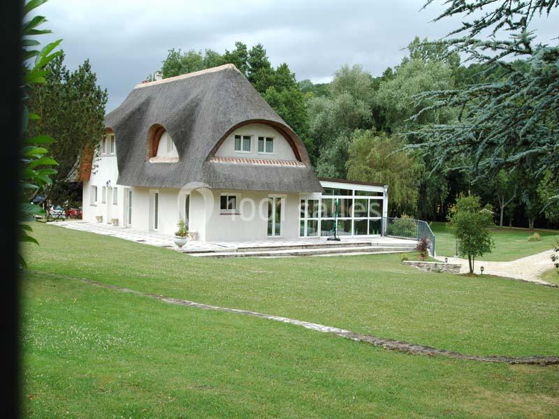 Maison au toit de chaume entourée d'un grand jardin verdoyant avec des arbres et une véranda attenante.