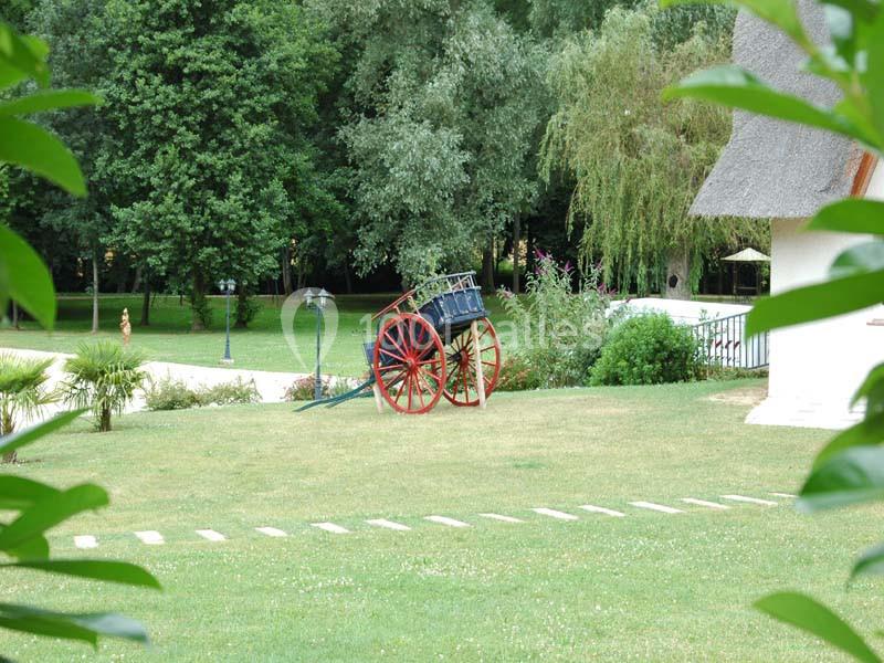 Charrette ancienne en bois rouge et bleu posée sur une pelouse, entourée d'arbres et de végétation.