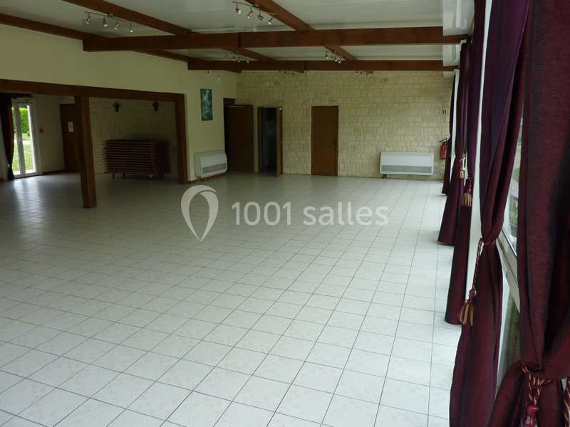 Salle vide avec sol carrelé, murs en pierre, poutres apparentes, grandes fenêtres avec rideaux bordeaux et portes en bois.