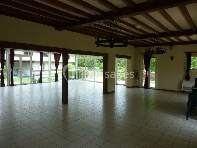 Salle spacieuse avec carrelage clair, poutres apparentes, grandes baies vitrées et rideaux bordeaux donnant sur un jardin.