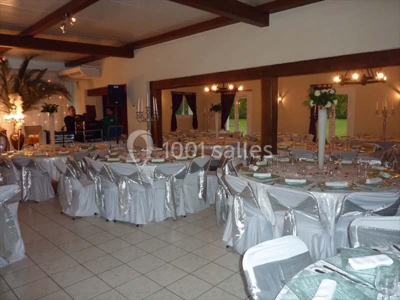 Salle de réception décorée avec des tables rondes dressées, nappes blanches et chaises couvertes, éclairage chaleureux.
