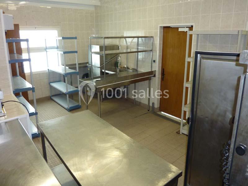 Cuisine professionnelle avec plans de travail en inox, étagères métalliques, évier et four, éclairée par une fenêtre.