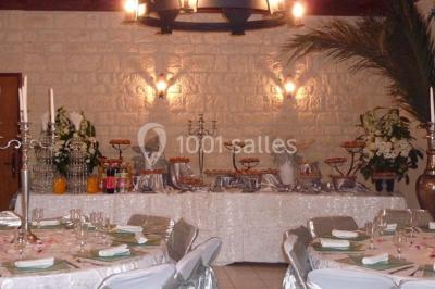 Salle décorée pour un événement avec tables dressées, chandeliers, nappes blanches et buffet garni en arrière-plan.