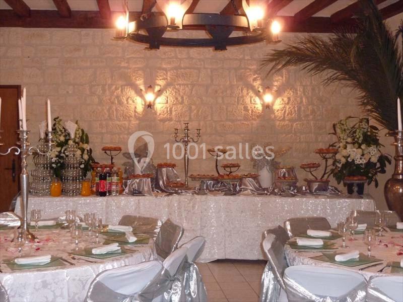 Salle décorée pour un événement avec tables dressées, chandeliers, nappes blanches et buffet garni en arrière-plan.