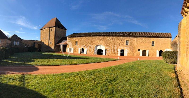 Bâtiment en pierre avec une tour et des arcades, entouré d'une pelouse et d'une allée sous un ciel bleu.