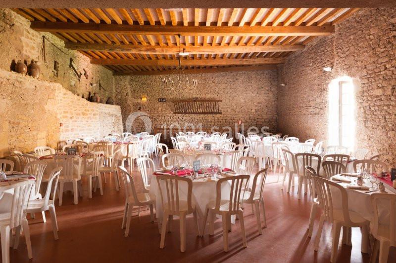 Salle en pierre avec tables rondes dressées, chaises blanches et lumière naturelle entrant par une grande fenêtre.