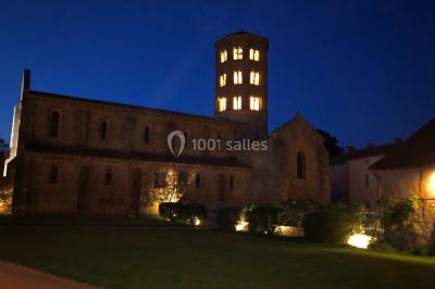 Église illuminée de nuit avec un clocher éclairé, vue depuis une allée avec des braseros allumés au premier plan.