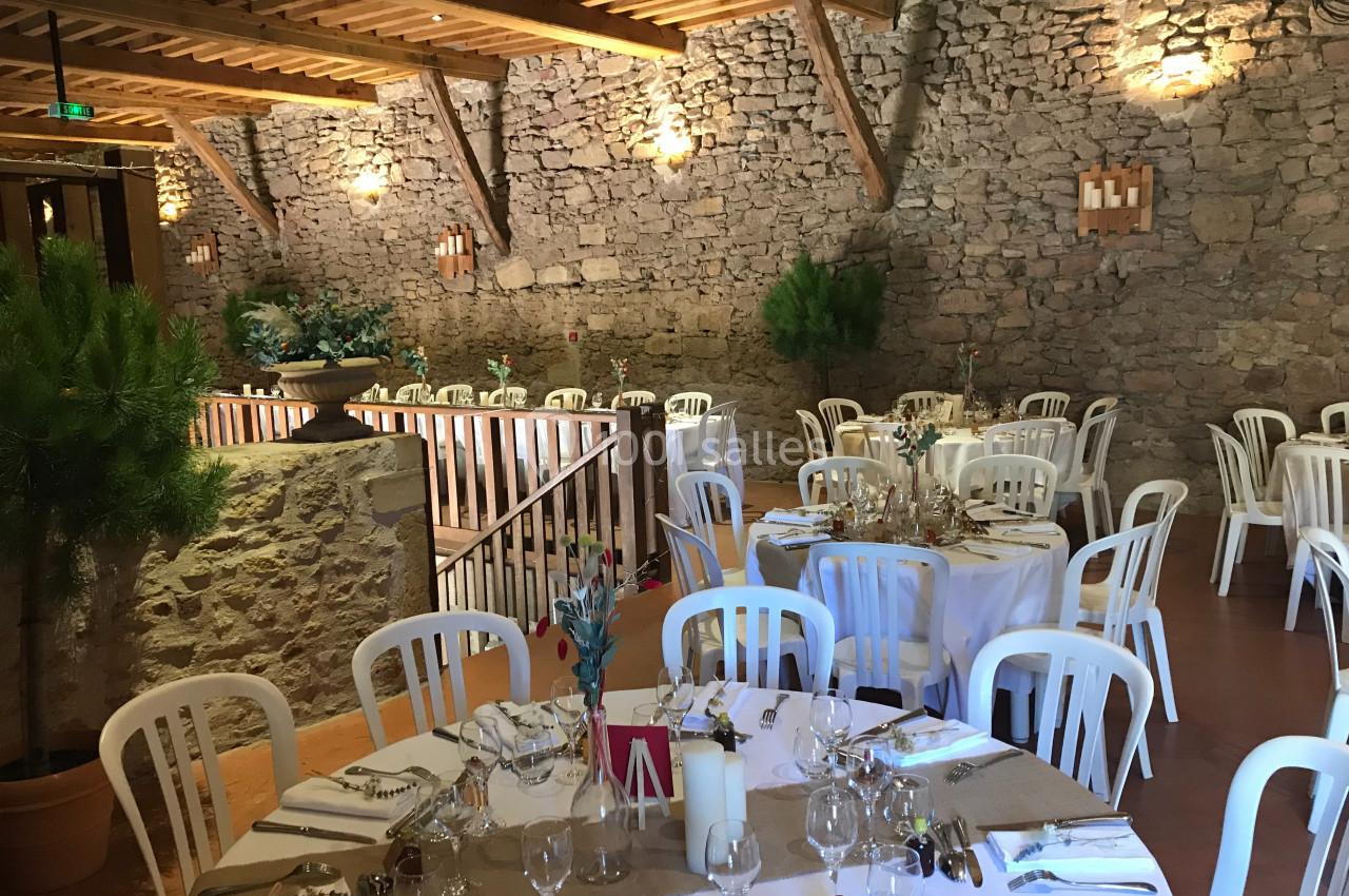 Salle de réception avec murs en pierre, tables rondes dressées avec nappes blanches et chaises en plastique.