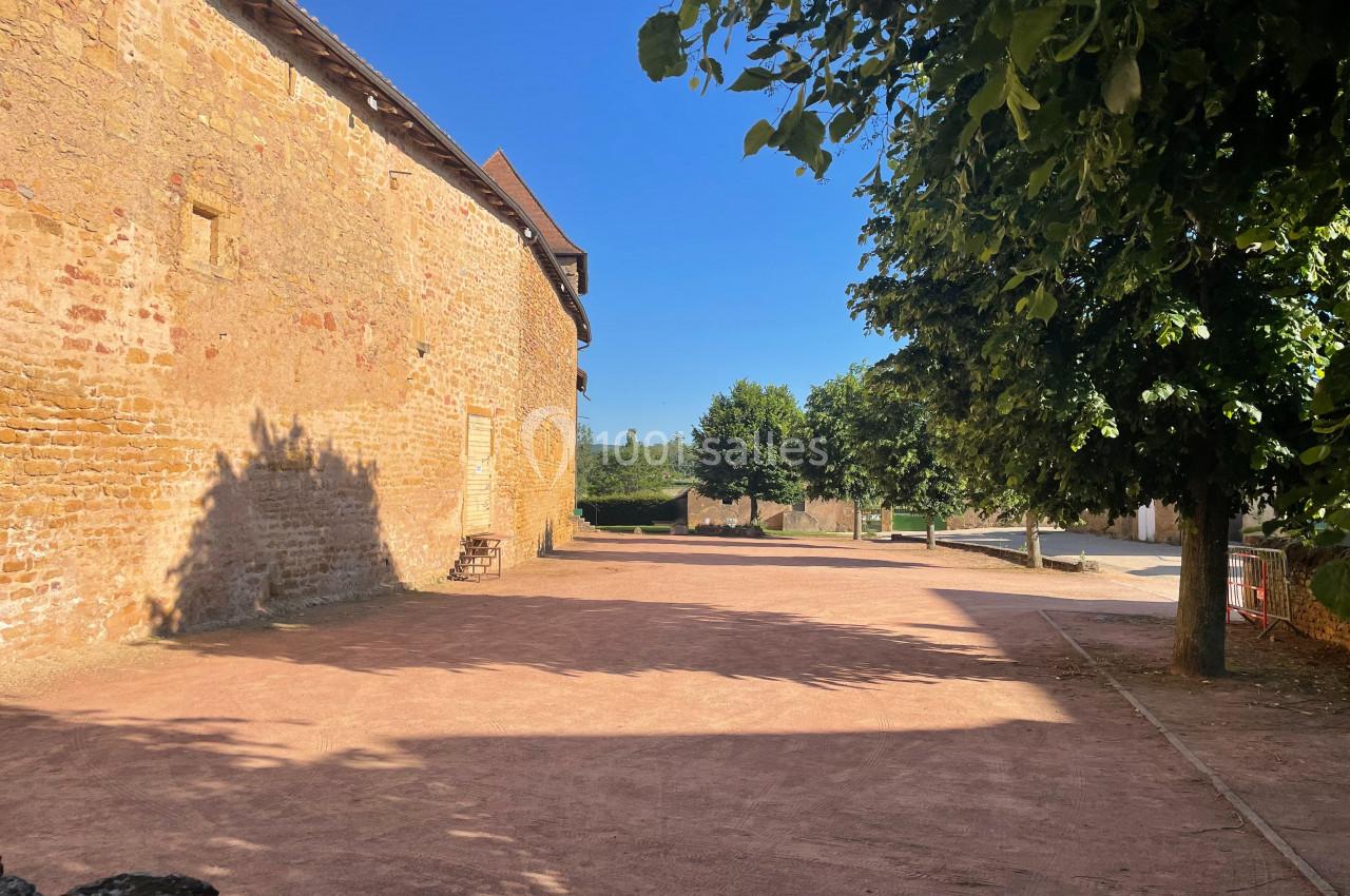 Cour en terre battue bordée d'un mur en pierre et d'arbres, sous un ciel bleu dégagé.