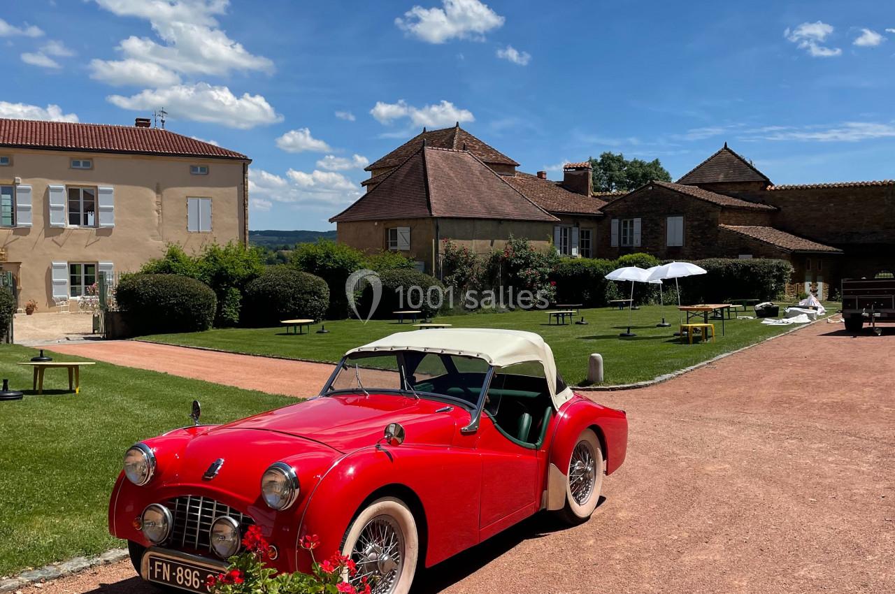 Voiture ancienne rouge décapotable stationnée devant un bâtiment en pierre avec pelouse et ciel dégagé.
