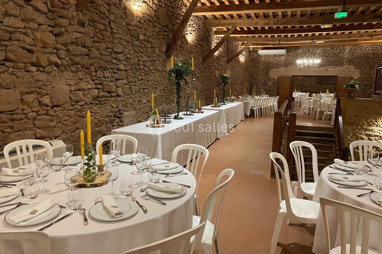 Salle en pierre aménagée pour un repas, avec tables rondes dressées et tables rectangulaires en fond.