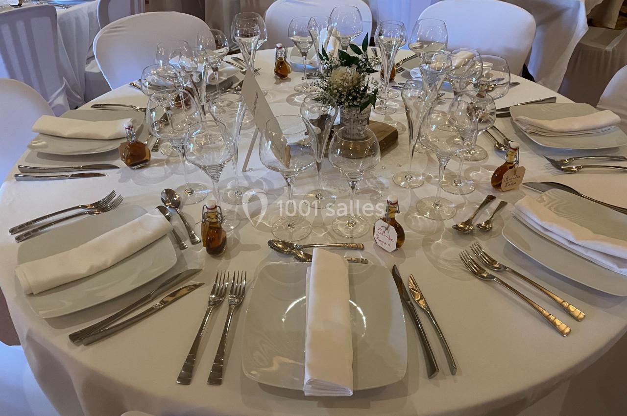 Table ronde élégamment dressée avec nappes blanches, couverts, verres, serviettes pliées et décoration florale centrale.
