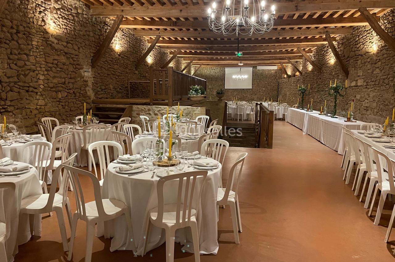 Salle de réception en pierre avec tables rondes dressées, chaises blanches et chandeliers, éclairée par un lustre central.